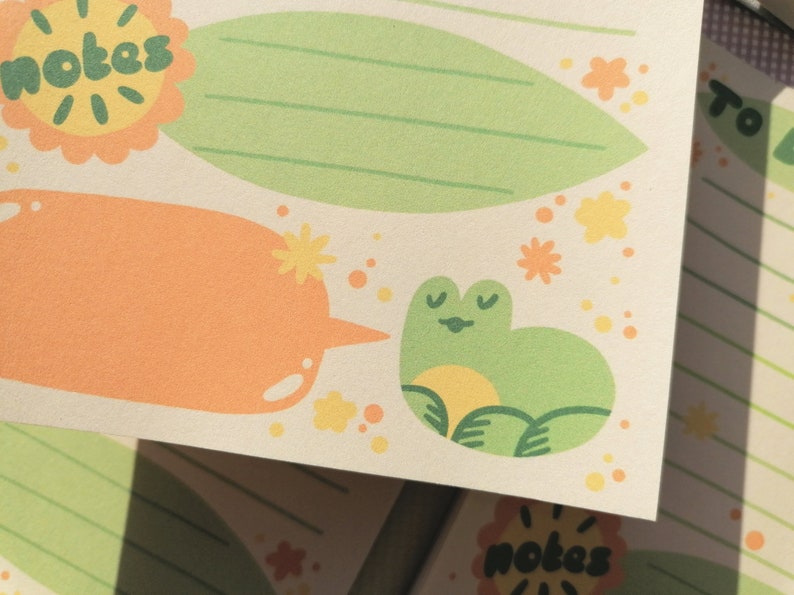 May include: A cream-colored notepad sheet featuring a green leaf-shaped section with horizontal lines, an orange speech bubble, and a small green frog illustration. The word notes is printed in dark green inside a yellow scalloped circle.