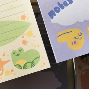 May include: Two sheets of stationery paper. The left sheet is cream-colored with green lines, a large green leaf, small yellow flowers, and a green frog illustration. The right sheet is light purple with the text notes, a white cloud, and yellow shapes.