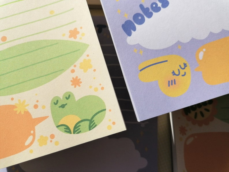 May include: Two sheets of stationery paper. The left sheet is cream-colored with green lines, a large green leaf, small yellow flowers, and a green frog illustration. The right sheet is light purple with the text notes, a white cloud, and yellow shapes.