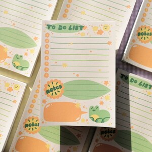 May include: A rectangular notepad featuring the text TO DO LIST at the top, with green horizontal lines for writing. The design includes orange flower borders, a green leaf shape labeled notes, an orange speech bubble, and a small green frog illustration.