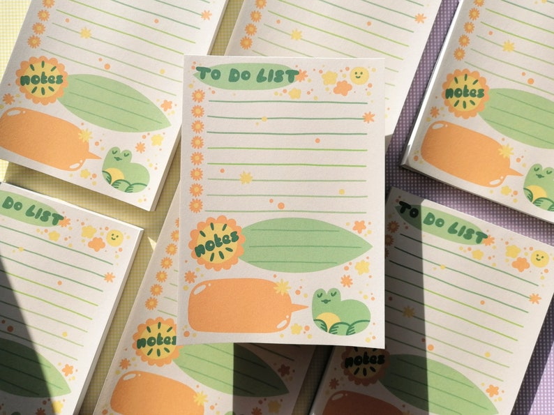 May include: A rectangular notepad featuring the text TO DO LIST at the top, with green horizontal lines for writing. The design includes orange flower borders, a green leaf shape labeled notes, an orange speech bubble, and a small green frog illustration.