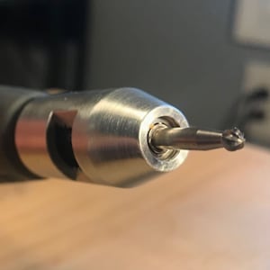 Puede incluir: Primer plano de una herramienta rotativa plateada con una pequeña fresa redonda en forma de diamante unida al extremo.