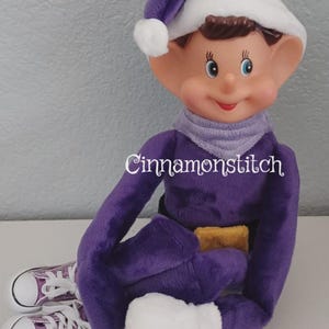 May include: A plush elf doll wearing a purple suit, a matching purple hat with a white trim and pom-pom, a light purple scarf, and white mittens. The doll wears purple canvas sneakers with white laces and soles. The text Cinnamonstitch is visible.