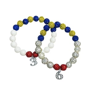 Könnte beinhalten: Zwei Perlenarmbänder mit weißen, blauen, gelben und roten Perlen. Die Armbänder haben silberne Anhänger mit den Zahlen 3 und 6.