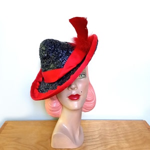 Può includere: Un cappello nero e rosso con una piuma rossa. Il cappello è fatto di paglia e ha un'ampia tesa. Il cappello è inclinato su un angolo sulla testa di un manichino.