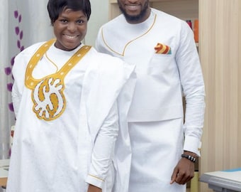 May include: A couple wearing white traditional African clothing. The woman's outfit features a gold embroidered design on the chest. The man's outfit has a gold trim and a pocket square with a colourful pattern. The couple are standing in front of a wooden shelf with spools of thread.