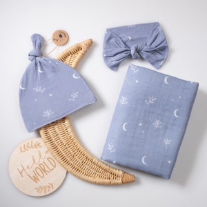 Puede incluir: Un conjunto de regalo para bebé con una manta de muselina azul, un gorro azul anudado, una diadema azul y un letrero de madera que dice "Hello World". La manta y el gorro tienen un patrón de luna y estrellas blancas.