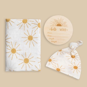 May include: A white baby blanket and hat with a gold sunburst pattern. A wooden round sign with the text "hello world" and blank lines for name, date, height, and weight.