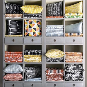 Puede incluir: Una estanter&iacute;a de madera gris con varios cajones y compartimentos llenos de cojines de tela con estampados coloridos y tela doblada. La tela presenta dise&ntilde;os geom&eacute;tricos y abstractos en negro, blanco, amarillo, naranja y gris.