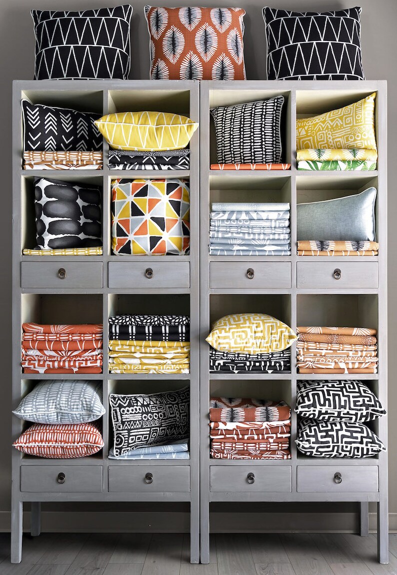 Puede incluir: Una estanter&iacute;a de madera gris con varios cajones y compartimentos llenos de cojines de tela con estampados coloridos y tela doblada. La tela presenta dise&ntilde;os geom&eacute;tricos y abstractos en negro, blanco, amarillo, naranja y gris.