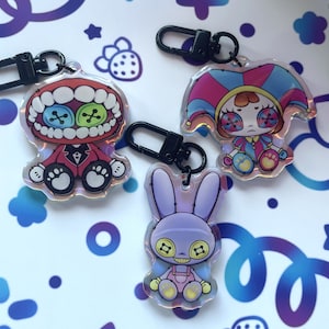 May include: Three colourful keychains featuring cartoon characters. The first keychain features a red and black character with a toothy grin and button eyes. The second keychain features a jester with a pink, blue and yellow costume. The third keychain features a purple bunny with a toothy grin and button eyes.