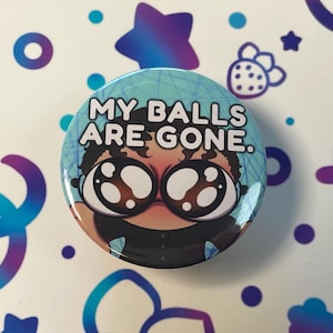 以下が含まれることがあります： 大きな目で漫画のキャラクターが描かれた青と白のボタンで、"MY BALLS ARE GONE."という文字が書かれています
