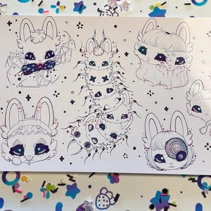 May include: A white art print featuring whimsical illustrations of bunny-like creatures and a centipede-like figure. The drawings are outlined in black, with details like bows and patterned eyes. The background is decorated with colorful confetti and star-shaped embellishments.