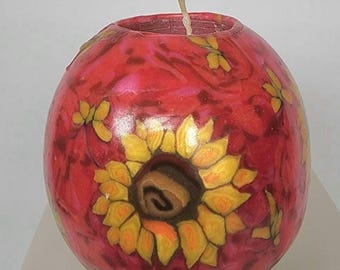 May include: A round, red candle with a sunflower design. The candle features a large sunflower in the center and smaller yellow flowers scattered across the surface. The candle has a wick and is set on a white surface.