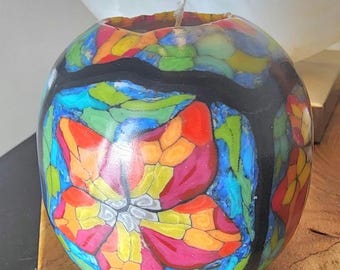 May include: A round, colorful candle with a floral design. The candle features a vibrant mosaic pattern in shades of red, orange, yellow, blue, and green, with a black border. A single wick sits atop the candle.