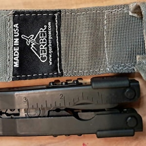 Puede incluir: Una herramienta multifunción negra con una regla integrada que muestra marcas de hasta 3 pulgadas, colocada debajo de una funda de nailon gris con una etiqueta de tela negra que dice MADE IN USA, GERBER y la dirección gerbergear.com en blanco.