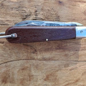 May include: A closed pocket knife featuring a dark brown textured handle with a wood-grain appearance. The knife includes a silver-toned metal bolster at one end and a metal bail or loop attached to the opposite end for carrying purposes.