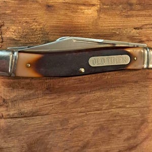 Op de afbeelding: Een inklapbaar zakmes met een gesloten handvat met een kleurverloop van donkerbruin naar crème. Het handvat bevat een metalen inlegstuk met de inscriptie OLD TIMER en gepolijste zilverkleurige metalen klinknagels aan beide uiteinden.