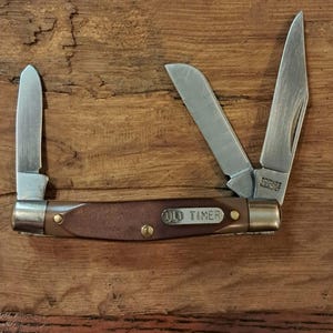 Può includere: Un coltellino tascabile pieghevole con tre lame in acciaio color argento estese. Il manico è marrone con una superficie testurizzata e presenta una placca intarsiata in metallo color argento incisa con il testo OLD TIMER. Rivetti in ottone.