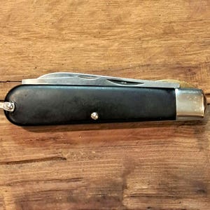 May include: A closed folding pocket knife with a smooth black handle, a silver-toned metal bolster at one end, and a small metal loop attached to the opposite end. The metallic blade is folded inside the handle, showing a thin silver edge.