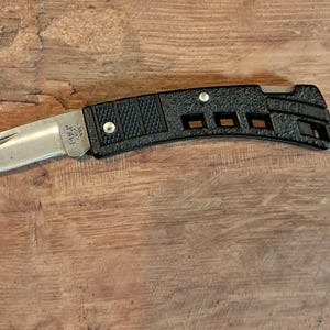May include: A folding pocket knife with a silver-colored metal blade and a black textured handle. The handle features three rectangular cutouts and the word BUCK embossed near the end. The blade is partially extended from the handle.