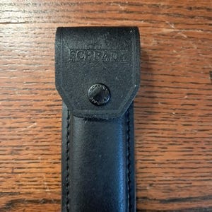May include: A black leather sheath with a rectangular flap and a snap button closure. The word SCHRADE is embossed on the front flap, and the word SCHRADE is also imprinted on the centre of the black metal snap button fastener.