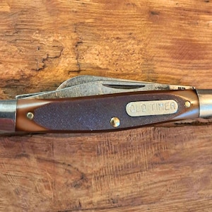 May include: A folding pocket knife with a brown handle featuring a textured grip and a silver-coloured metal bolster at each end. The handle includes an embedded silver-coloured plate with the text OLD TIMER. The metal blade is partially visible.