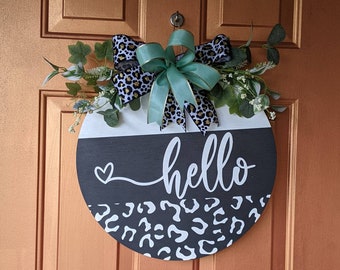 May include: A round wooden door hanger with a black and white leopard print design. The top half of the hanger is white and the bottom half is black. The word "hello" is written in white cursive script on the black portion of the hanger. A small white heart is above the word "hello". The hanger is decorated with a green and black leopard print bow and green foliage.