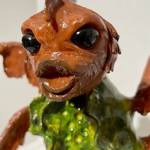 May include: A detailed sculpture of a creature with a brown head, large black eyes, and a smiling mouth. The creature wears a green and yellow textured garment. The sculpture has a glossy finish and is set against a plain background.