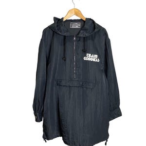 Puede incluir: Chaqueta anorak negra con capucha, cierre de media cremallera y bolsillo delantero. La chaqueta tiene el texto blanco "TEAM GODDESS". Cuelga de una percha de madera.
