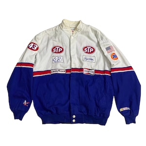 May include: A white and blue racing jacket with red accents. The jacket has several patches, including "STP", "43", "GoodYear", "Champion", and "NASCAR".