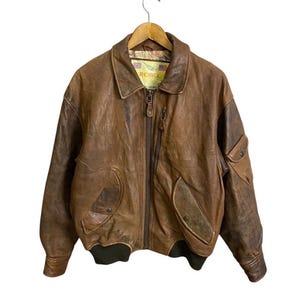 Può includere: Una giacca bomber in pelle marrone con chiusura a zip e due tasche. La giacca ha un'etichetta con la scritta "ROBOCOP" sul davanti.