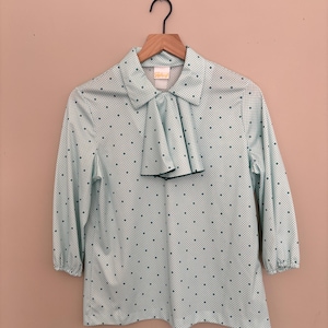 Puede incluir: Una blusa verde claro con estampado de lunares blancos y una pajarita verde. La blusa tiene cuello y mangas tres cuartos.