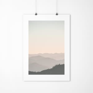 Op de afbeelding: Een ingelijste foto van een bergketen met een zachte, wazige roze en blauwe gradiënt in de lucht. De bergen zijn gesilhouetteerd tegen de lucht, met een paar bomen zichtbaar op de voorgrond.