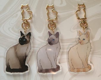 May include: Three keychains featuring different coloured Siamese cats. Each keychain has a gold metal cat-shaped charm attached to a clear acrylic charm with a cat illustration.