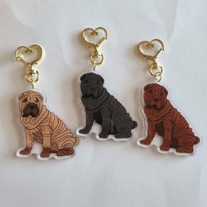 May include: Three Shar Pei dog keychains in tan, black, and brown. Each keychain features a gold-colored heart-shaped clasp. The dogs are sitting, with detailed wrinkles and a clear acrylic backing.