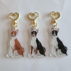 May include: Three Basenji dog keychains with gold-colored heart-shaped clasps. The dogs are illustrated in brown, black, and white, each in a sitting position. The keychains are made of clear acrylic.