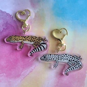 Może przedstawiać: Dwa breloczki do kluczy z motywami gekona lamparciego. Jeden breloczek ma żółtego, czarnego i białego gekona ze złotym zapięciem w kształcie serca. Drugi breloczek ma czarno-białego gekona ze złotym zapięciem w kształcie serca.