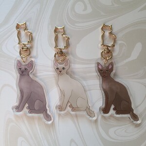 May include: Three clear acrylic keyrings featuring a cartoon illustration of a cat in three different colours: brown, white, and grey. Each keyring has a gold metal ring and a gold metal clasp.