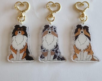 May include: Three dog-themed keychains with gold-colored heart-shaped clasps. The dog illustrations feature various coat colors: black, brown, white, and gray. The keychains are made of clear acrylic.