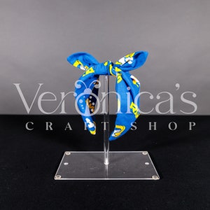 Pode incluir: Faixa de cabeça azul com estampa amarela e branca. A faixa de cabeça está amarrada em um laço e está em um suporte transparente. Veronica's Craftshop está impresso no suporte.