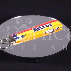 May include: A yellow, red, and blue lanyard with the word "Astros" in white lettering. The lanyard has a silver key ring attached. The product is displayed on a reflective surface.