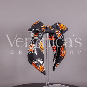 May include: A black and orange Cincinnati Bengals bow. The bow features a repeating pattern of the team's logo and helmet. The bow is displayed on a clear stand against a grey background. The text "Veronica's Craft Shop" is in the background.