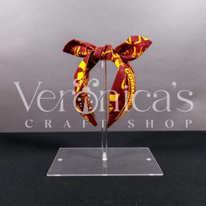 May include: Red and gold headband with a bow, featuring a repeating pattern of the words "Commonwealth" and "Community".