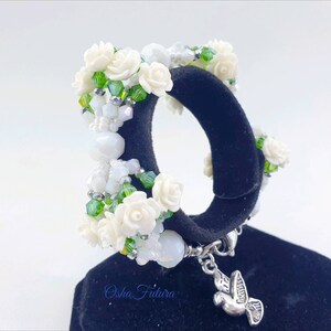 Puede incluir: Pulsera de flores blancas con cuentas verdes y un dije de plata en forma de paloma. La pulsera est&aacute; en un expositor negro. El texto "Osha Futura" es visible en el expositor.
