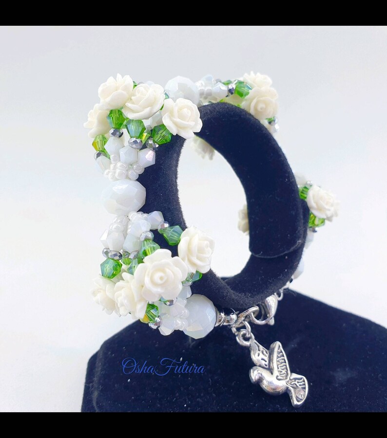 Puede incluir: Pulsera de flores blancas con cuentas verdes y un dije de plata en forma de paloma. La pulsera est&aacute; en un expositor negro. El texto "Osha Futura" es visible en el expositor.