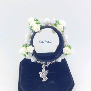 Puede incluir: Pulsera de cuentas blancas con flores blancas y hojas verdes. La pulsera tiene un dije de paloma plateada. La pulsera est&aacute; en un expositor negro. "Osha Futura" est&aacute; escrito en el expositor.