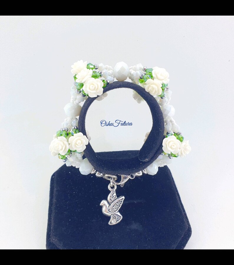 Puede incluir: Pulsera de cuentas blancas con flores blancas y hojas verdes. La pulsera tiene un dije de paloma plateada. La pulsera est&aacute; en un expositor negro. "Osha Futura" est&aacute; escrito en el expositor.
