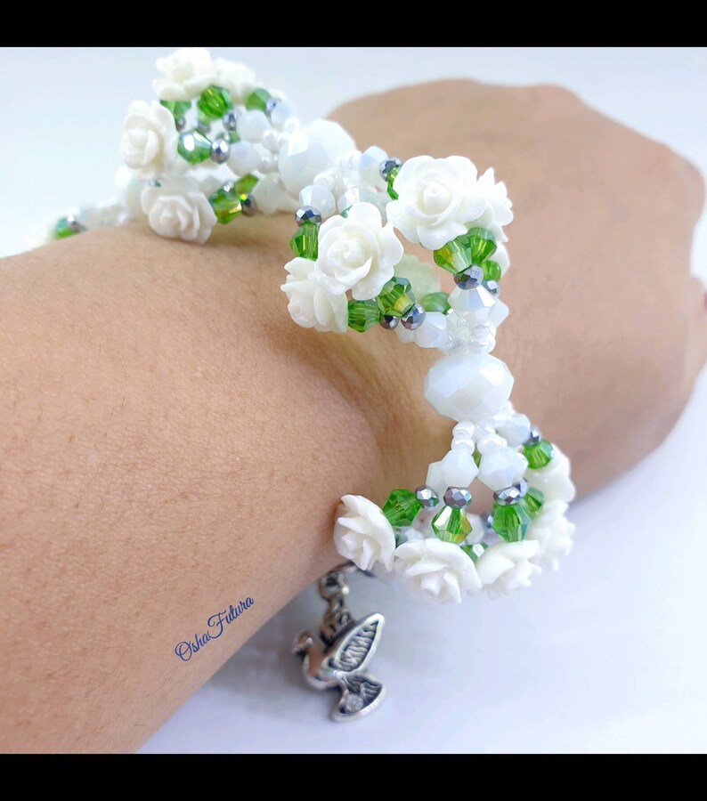 Puede incluir: Pulsera de cuentas blancas con cuentas verdes y plateadas, con detalles de flores blancas y un dije de paloma plateada.