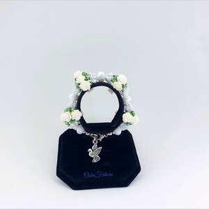 Puede incluir: Pulsera blanca de cuentas con flores blancas y hojas verdes. La pulsera tiene un dije de paloma plateada y el texto "Osha Futura" en un expositor de terciopelo negro.